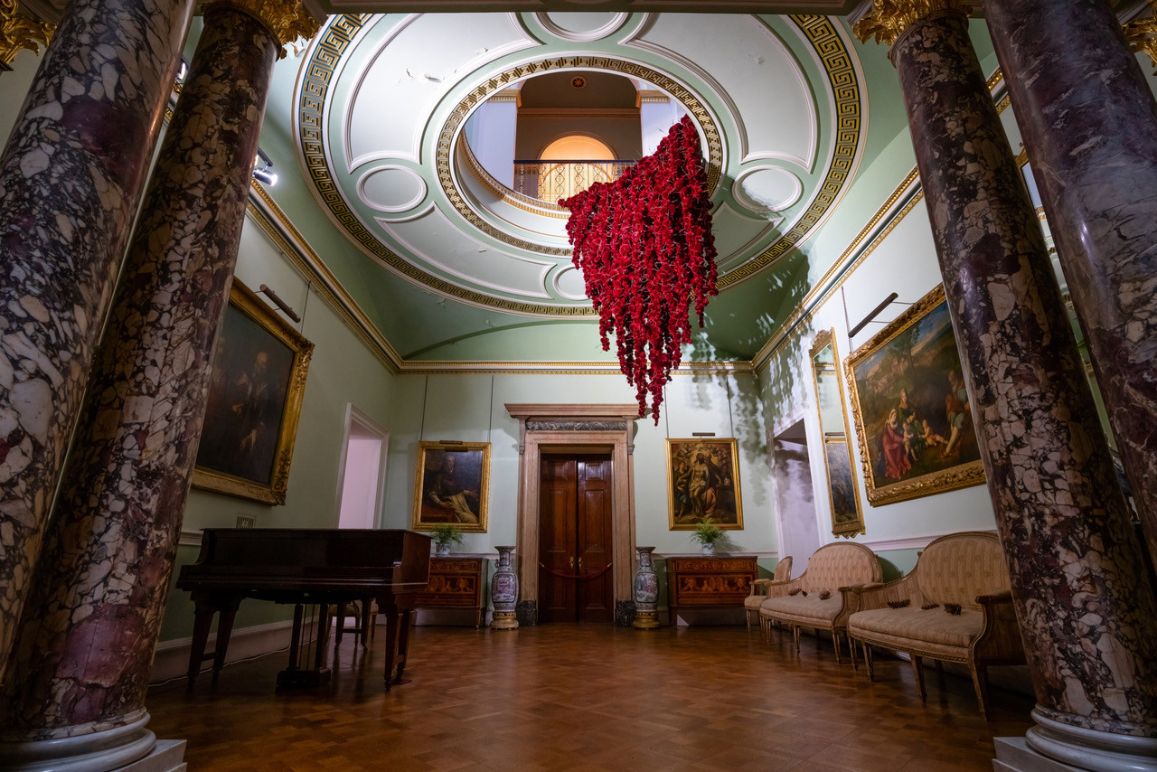Gathering
site specific installation
Tatton Park Mansion 2019
silk velvet, reclaimed military barbed wire
Susie MacMurray has an uncanny knack of going to the heart of the matter. Her work is somehow immediately evocative, reeling in the viewer to contemplate a sense of place, a feeling of loss, and a desire to touch. Her unusual combinations of materials and subject matter, in site-specific installations, are always intriguing and poetic. They provoke an interaction with the viewer, which draws on both physical and cultural associations.
In 'Gathering,' designed specifically to hang from the circular piercing under the rotunda of the staircase hall at Tatton Park, MacMurray returns to her constant theme of the ephemeral nature of life. To create this installation, she has worked regularly with small groups of volunteers, to hand sew tiny, luxurious, red velvet, ball-like elements, which she has then assembled to cascade in different configurations from the edge of the opening. The overall effect is of an eerie, dripping canopy of red velvet Spanish moss.
The luxury and softness of the deep red silk velvet, is impaled by protruding lengths of barbed wire, used by the MoD to train soldiers going to the Falklands, Kosovo and Afghanistan. The use of a material associated with conflict can be read as alluding to the narrative of cancer as a battle. This bringing together of such diametrically opposed materials lies at the core of MacMurray's approach. She enjoys "the conversations between not always expected combinations of materials, or materials in contexts in which you are not expecting to see them." Such juxtapositions are simultaneously appealing and disturbing. MacMurray works with and through materials, using them as a way of thinking about and expressing more conceptual ideas.
MacMurray is no stranger to loss; her husband died very young and very rapidly of cancer. In 'Gathering,' she examines this experience. The red silk she perceives as sensual, positive, bodily and precious, expressing love and desire and the living of life to the full. Yet the velvet impaled by the barbed wire is strangely beautiful and evocative. "Your life," she explains, "gets pierced by this sudden ordeal." The hospice helps by gathering people in its fold. Indeed many of the volunteers involved in the making of 'Gathering,' have been helped by the Hospice in this enfolding, supportive way, which MacMurray expresses in the gathering together of the red velvet elements.
MacMurray uses all manner of unlikely objects from rubber tubing, to barbed wire, to hair and wax, via a range of textiles and cords, combining them in strange assortments, to make them things of beauty and resonance. Her work is both solid and ethereal. She looks at the ephemeral nature of life. At life and loss, the soft, imperfect, human body that despite its imperfections is so glorious. She entices the visitor into contemplating the fragility of life, but also its joys.
Corrine Julius
June 2019
Susie MacMurray was invited to create this work to both celebrate and highlight the 20th anniversary of the Art Fair, which was established in 1999 to raise funds for the East Cheshire Hospice.
To purchase a piece of Gathering visit <a href='https://www.gatheringattatton.com/store'>gatheringattatton</a>
All funds raised go to East Cheshire Hospice.