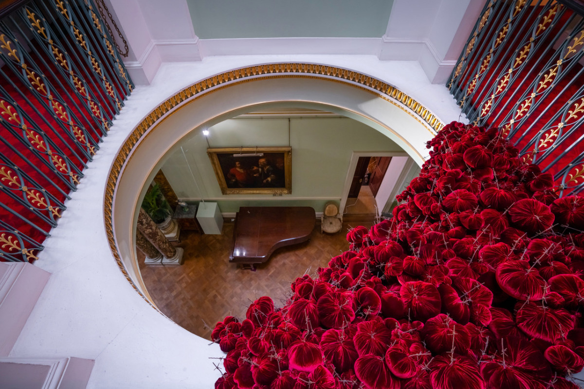 Gathering
site specific installation
Tatton Park Mansion 2019
silk velvet, reclaimed military barbed wire
Susie MacMurray has an uncanny knack of going to the heart of the matter. Her work is somehow immediately evocative, reeling in the viewer to contemplate a sense of place, a feeling of loss, and a desire to touch. Her unusual combinations of materials and subject matter, in site-specific installations, are always intriguing and poetic. They provoke an interaction with the viewer, which draws on both physical and cultural associations.
In 'Gathering,' designed specifically to hang from the circular piercing under the rotunda of the staircase hall at Tatton Park, MacMurray returns to her constant theme of the ephemeral nature of life. To create this installation, she has worked regularly with small groups of volunteers, to hand sew tiny, luxurious, red velvet, ball-like elements, which she has then assembled to cascade in different configurations from the edge of the opening. The overall effect is of an eerie, dripping canopy of red velvet Spanish moss.
The luxury and softness of the deep red silk velvet, is impaled by protruding lengths of barbed wire, used by the MoD to train soldiers going to the Falklands, Kosovo and Afghanistan. The use of a material associated with conflict can be read as alluding to the narrative of cancer as a battle. This bringing together of such diametrically opposed materials lies at the core of MacMurray's approach. She enjoys "the conversations between not always expected combinations of materials, or materials in contexts in which you are not expecting to see them." Such juxtapositions are simultaneously appealing and disturbing. MacMurray works with and through materials, using them as a way of thinking about and expressing more conceptual ideas.
MacMurray is no stranger to loss; her husband died very young and very rapidly of cancer. In 'Gathering,' she examines this experience. The red silk she perceives as sensual, positive, bodily and precious, expressing love and desire and the living of life to the full. Yet the velvet impaled by the barbed wire is strangely beautiful and evocative. "Your life," she explains, "gets pierced by this sudden ordeal." The hospice helps by gathering people in its fold. Indeed many of the volunteers involved in the making of 'Gathering,' have been helped by the Hospice in this enfolding, supportive way, which MacMurray expresses in the gathering together of the red velvet elements.
MacMurray uses all manner of unlikely objects from rubber tubing, to barbed wire, to hair and wax, via a range of textiles and cords, combining them in strange assortments, to make them things of beauty and resonance. Her work is both solid and ethereal. She looks at the ephemeral nature of life. At life and loss, the soft, imperfect, human body that despite its imperfections is so glorious. She entices the visitor into contemplating the fragility of life, but also its joys.
Corrine Julius
June 2019
Susie MacMurray was invited to create this work to both celebrate and highlight the 20th anniversary of the Art Fair, which was established in 1999 to raise funds for the East Cheshire Hospice.
To purchase a piece of Gathering visit <a href='https://www.gatheringattatton.com/store'>gatheringattatton</a>
All funds raised go to East Cheshire Hospice.