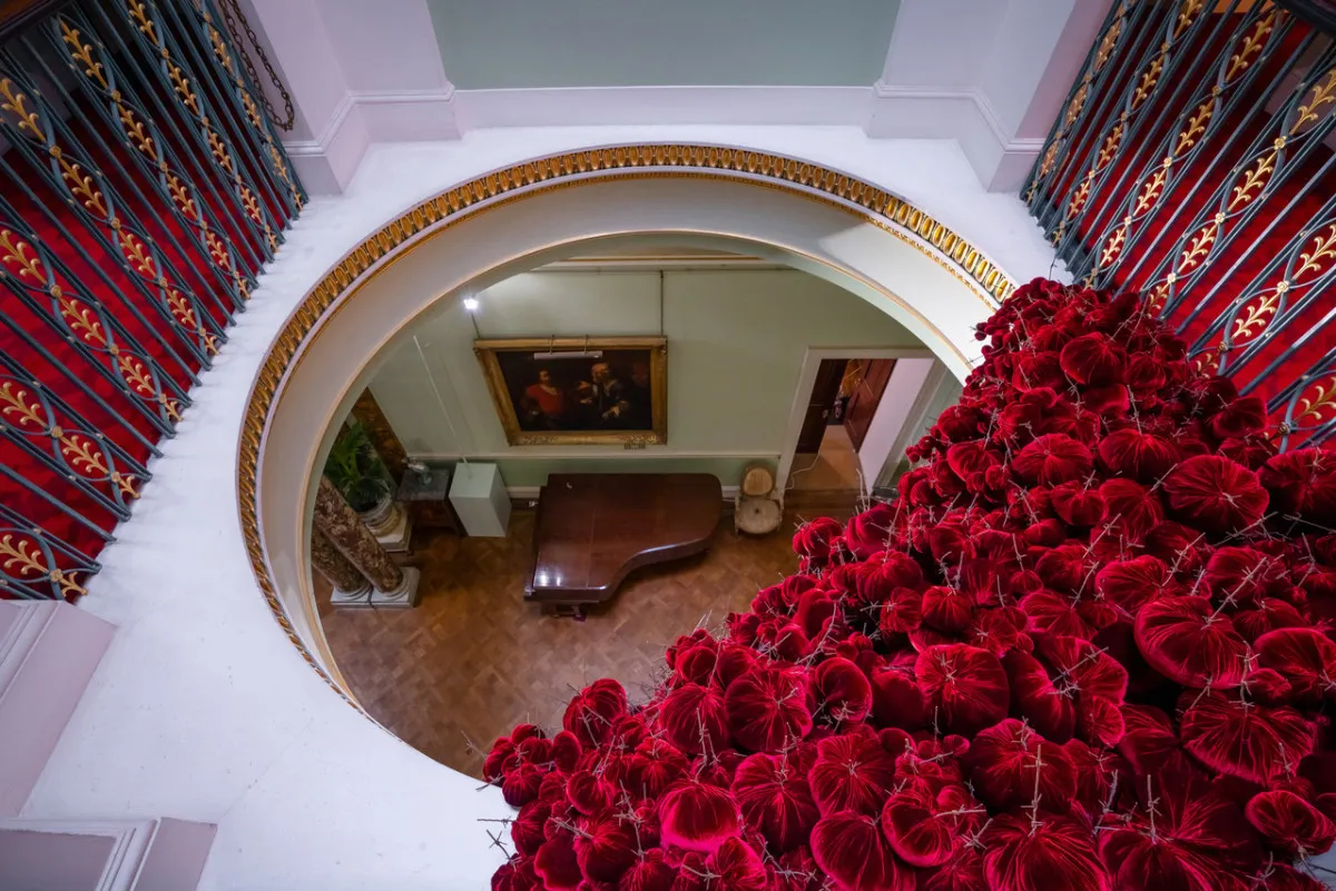 Gathering
site specific installation
Tatton Park Mansion 2019
silk velvet, reclaimed military barbed wire
Susie MacMurray has an uncanny knack of going to the heart of the matter. Her work is somehow immediately evocative, reeling in the viewer to contemplate a sense of place, a feeling of loss, and a desire to touch. Her unusual combinations of materials and subject matter, in site-specific installations, are always intriguing and poetic. They provoke an interaction with the viewer, which draws on both physical and cultural associations.
In 'Gathering,' designed specifically to hang from the circular piercing under the rotunda of the staircase hall at Tatton Park, MacMurray returns to her constant theme of the ephemeral nature of life. To create this installation, she has worked regularly with small groups of volunteers, to hand sew tiny, luxurious, red velvet, ball-like elements, which she has then assembled to cascade in different configurations from the edge of the opening. The overall effect is of an eerie, dripping canopy of red velvet Spanish moss.
The luxury and softness of the deep red silk velvet, is impaled by protruding lengths of barbed wire, used by the MoD to train soldiers going to the Falklands, Kosovo and Afghanistan. The use of a material associated with conflict can be read as alluding to the narrative of cancer as a battle. This bringing together of such diametrically opposed materials lies at the core of MacMurray's approach. She enjoys "the conversations between not always expected combinations of materials, or materials in contexts in which you are not expecting to see them." Such juxtapositions are simultaneously appealing and disturbing. MacMurray works with and through materials, using them as a way of thinking about and expressing more conceptual ideas.
MacMurray is no stranger to loss; her husband died very young and very rapidly of cancer. In 'Gathering,' she examines this experience. The red silk she perceives as sensual, positive, bodily and precious, expressing love and desire and the living of life to the full. Yet the velvet impaled by the barbed wire is strangely beautiful and evocative. "Your life," she explains, "gets pierced by this sudden ordeal." The hospice helps by gathering people in its fold. Indeed many of the volunteers involved in the making of 'Gathering,' have been helped by the Hospice in this enfolding, supportive way, which MacMurray expresses in the gathering together of the red velvet elements.
MacMurray uses all manner of unlikely objects from rubber tubing, to barbed wire, to hair and wax, via a range of textiles and cords, combining them in strange assortments, to make them things of beauty and resonance. Her work is both solid and ethereal. She looks at the ephemeral nature of life. At life and loss, the soft, imperfect, human body that despite its imperfections is so glorious. She entices the visitor into contemplating the fragility of life, but also its joys.
Corrine Julius
June 2019
Susie MacMurray was invited to create this work to both celebrate and highlight the 20th anniversary of the Art Fair, which was established in 1999 to raise funds for the East Cheshire Hospice.
To purchase a piece of Gathering visit <a href='https://www.gatheringattatton.com/store'>gatheringattatton</a>
All funds raised go to East Cheshire Hospice.